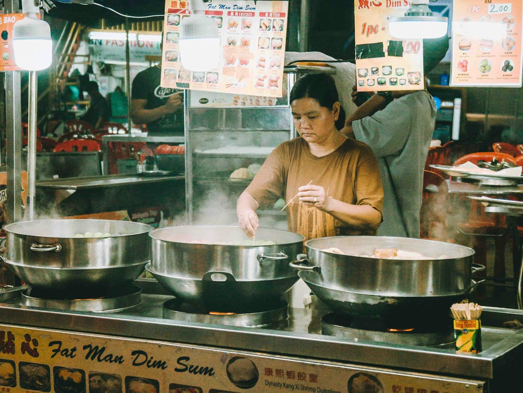 An Asian woman cooking dim sum at a vibrant street food stall in Kuala Lumpur.